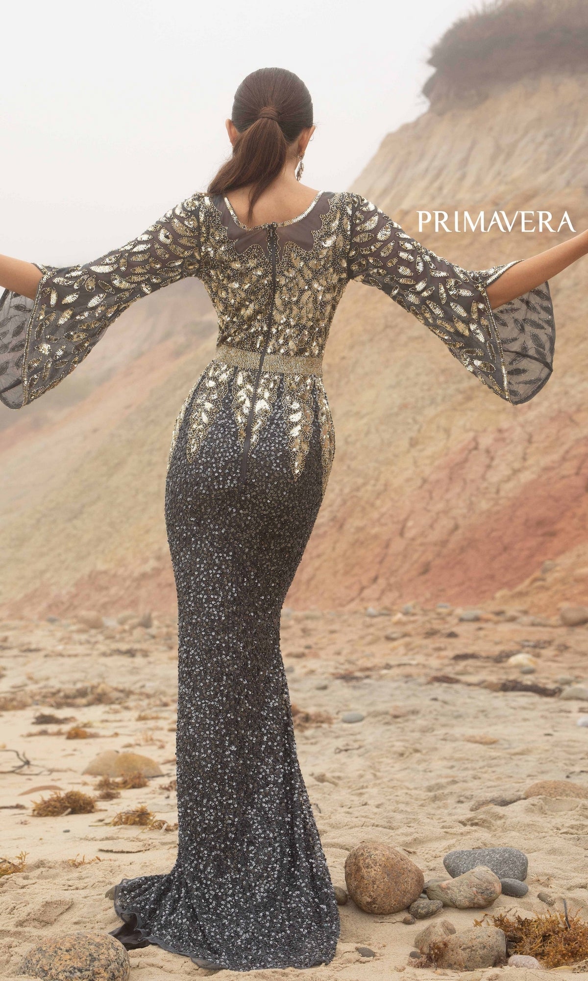 A woman wearing the Bell-Sleeve Long Beaded Evening Gown 1424, featuring flared sleeves and a sequin skirt, stands on a sandy beach facing away.